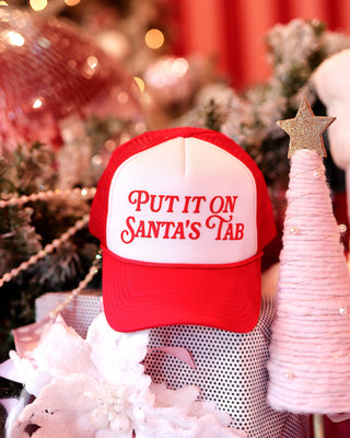 Red and white cap with 'Put it on Santa's Tab' text in a festive setting with Christmas decorations.