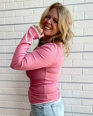 A woman posing in front of a white brick wall, wearing a fitted pink long-sleeve top with lace trim on the cuffs and a light blue denim mini skirt.