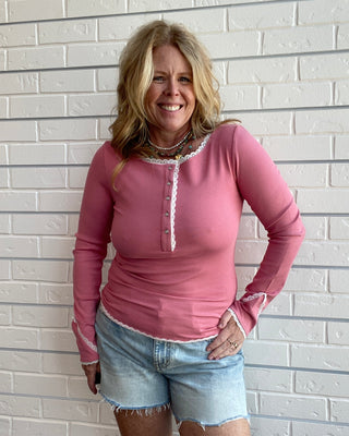 A smiling blonde woman wearing a pink long-sleeve henley top with white lace trim and light blue denim shorts, standing against a white brick wall.