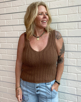 Woman wearing a brown tank top and light blue jeans standing against a light brick wall.