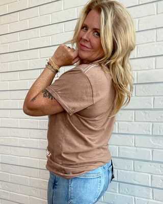 A woman with blonde hair wearing a light brown short-sleeve t-shirt and light blue jeans, posing against a white brick wall.