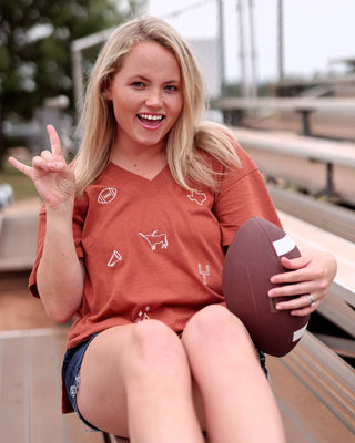 A smiling blonde woman sitting on outdoor bleachers holds a football and wears a rust-orange v-neck t-shirt with small white football-themed graphics, paired with denim shorts.