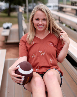 A smiling blonde woman sits on outdoor bleachers holding a football, wearing a rust-orange v-neck t-shirt with small white graphic icons and dark denim shorts.