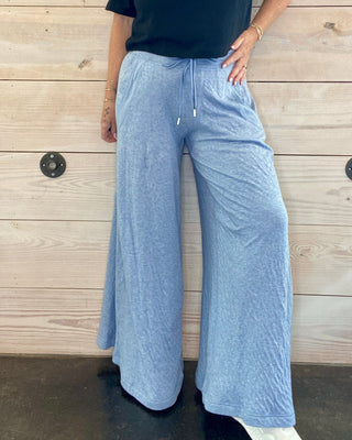 Person wearing a black t-shirt and light blue wide-leg pants standing against a wooden wall.