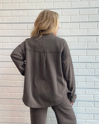 A woman stands with her back to the camera wearing a matching dark brown lounge set consisting of a long-sleeve button-up shirt and wide-leg pants, set against a white brick wall.