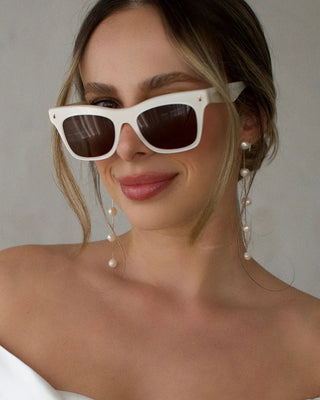 Woman wearing white sunglasses and pearl earrings against a neutral background