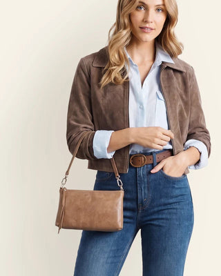 A woman models a brown suede jacket, blue jeans, a light blue button-down shirt, and a brown leather crossbody bag with a matching belt, set against a plain cream background.