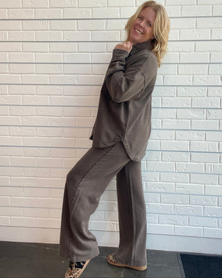 A woman posing in a matching dark brown lounge set consisting of a long-sleeve button-up shirt and wide-leg trousers, standing against a white brick wall.