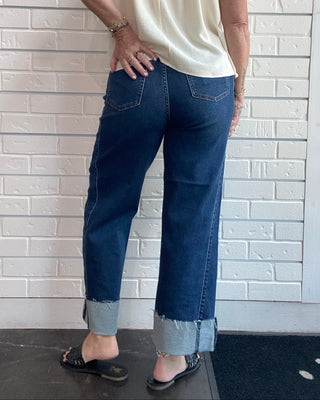 A woman shown from the back wearing dark blue cuffed jeans and a cream sleeveless top, standing against a white brick wall.