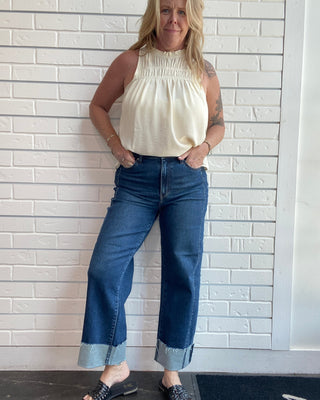 A woman modeling a cream sleeveless smocked top and dark blue cropped wide-leg jeans with rolled cuffs, standing against a white brick wall.