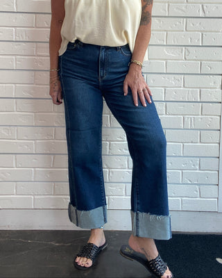 A woman modeling a cream sleeveless smocked blouse and dark blue cropped jeans with rolled hems, standing against a white brick wall.