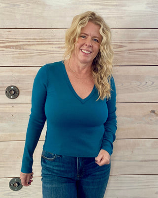 Woman wearing a teal long-sleeve top and jeans standing against a wooden wall.