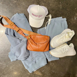 Light blue sweatpants, brown leather bag, white sneakers, and a white cap with 'marge' text on a gray concrete floor.