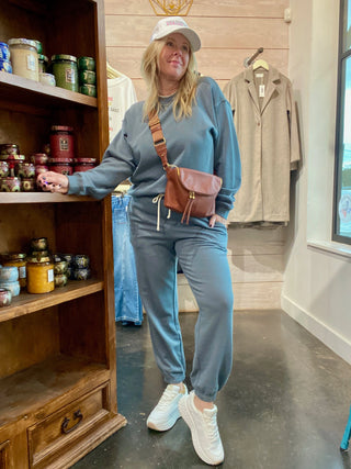Woman in a blue tracksuit with a brown bag standing in a room with wooden shelves and clothing.