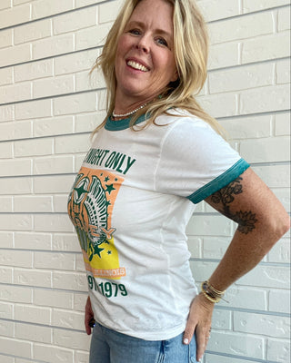 A woman wearing a white ringer t-shirt with teal and orange graphics, paired with light blue jeans, standing against a white brick wall.