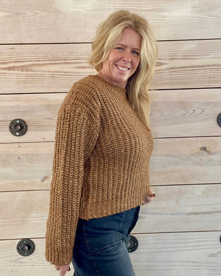 Woman wearing a brown sweater and blue jeans standing against a wooden wall.