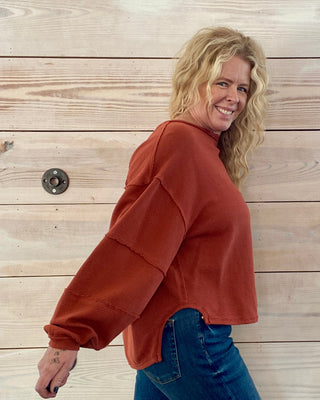Woman wearing a rust-colored sweater and blue jeans standing against a wooden wall.