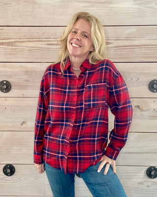 Woman wearing a red plaid shirt and blue jeans standing against a wooden wall.