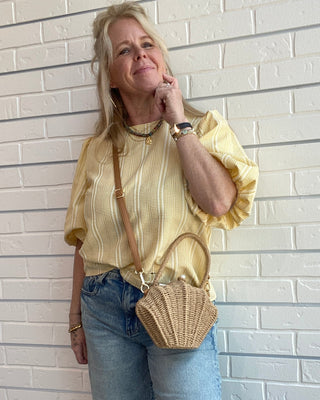 Woman with a straw shell crosbody bag standing against a white brick wall.