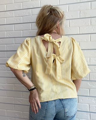Person wearing a yellow blouse with a bow detail on the back, standing against a light brick wall.