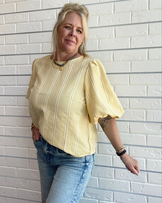 Woman wearing a yellow blouse and blue jeans against a white brick wall.