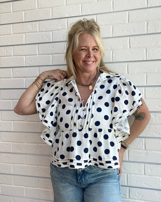 Woman wearing a polka dot blouse and jeans against a white brick wall.