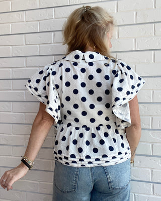 Person wearing a white blouse with black polka dots and blue jeans against a light brick wall.