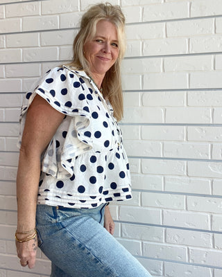 Woman wearing a polka dot blouse and jeans against a white brick wall.