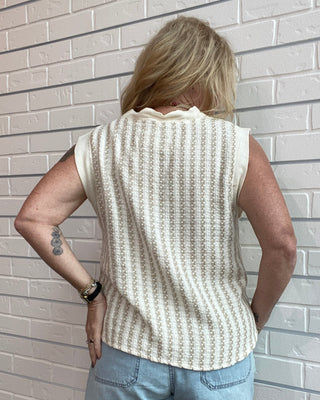 Person wearing a sleeveless patterned top and denim shorts against a light brick wall.