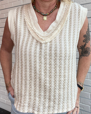 Person wearing a sleeveless beige top with lace details against a tiled wall.