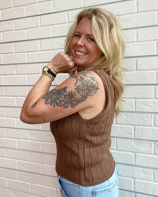 Woman with a tattoo on her arm wearing a brown sleeveless top and blue jeans against a light brick wall.