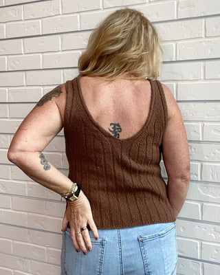 Person wearing a brown tank top and blue jeans standing against a light brick wall.