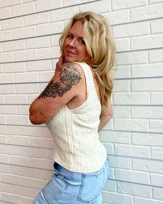 Woman with a tattoo on her arm wearing a white sleeveless top and blue jeans against a light brick wall.
