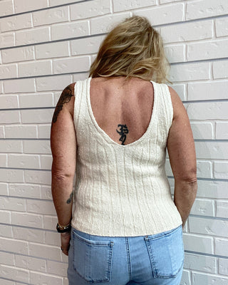 Person wearing a white sleeveless top and blue jeans against a light brick wall.