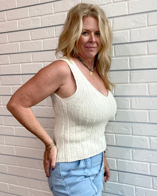 Woman wearing a white sleeveless top and light blue jeans against a light brick wall.