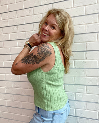 Woman wearing a green sleeveless top and blue jeans against a white brick wall.