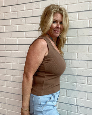 Woman wearing a brown sleeveless top and light blue jeans standing against a white brick wall.