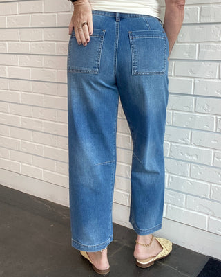 Person wearing a white sleeveless top and blue jeans standing against a light brick wall.