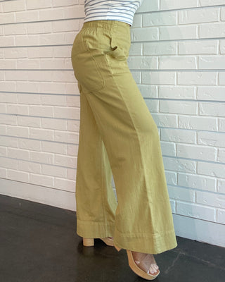 Woman wearing a striped tank top and green wide-leg pants against a white brick wall.