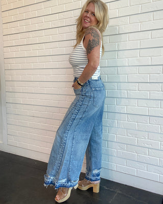 Woman wearing a striped tank top and wide-leg jeans against a white brick wall.