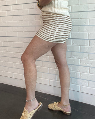 Woman in a white sleeveless top and striped skirt standing against a light brick wall.