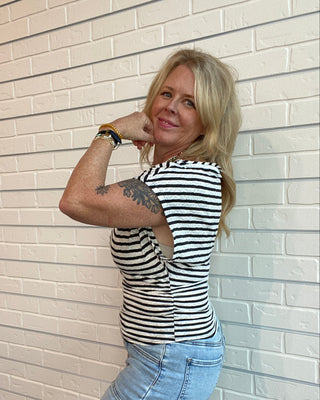 Woman in a striped shirt and jeans leaning against a light-colored brick wall.