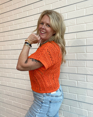 Woman wearing an orange knitted top and blue jeans standing against a light brick wall.