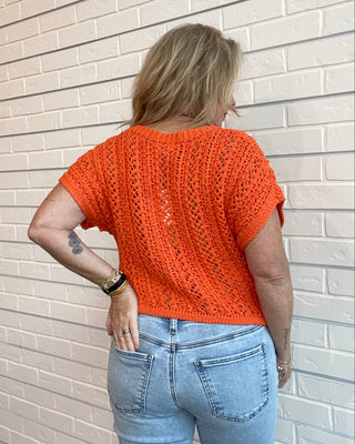 Person wearing an orange crochet top and blue jeans standing against a white brick wall.