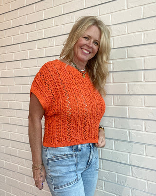 Woman wearing an orange knitted top and blue jeans standing against a light brick wall.