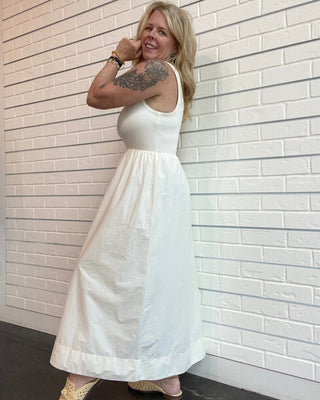 Woman in a white dress standing against a light brick wall.