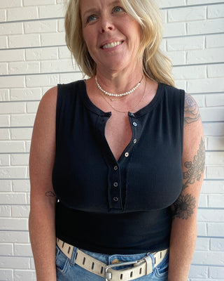 Woman wearing a black sleeveless top and blue jeans against a white brick wall.