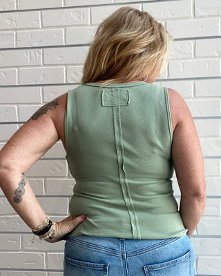 Person wearing a green sleeveless top and blue jeans against a white brick wall.