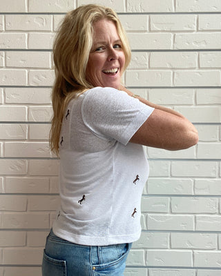 Woman wearing a white t-shirt with small graphic designs, standing against a light brick wall.