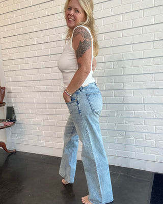 Woman wearing a white tank top and light blue jeans standing against a white brick wall.
